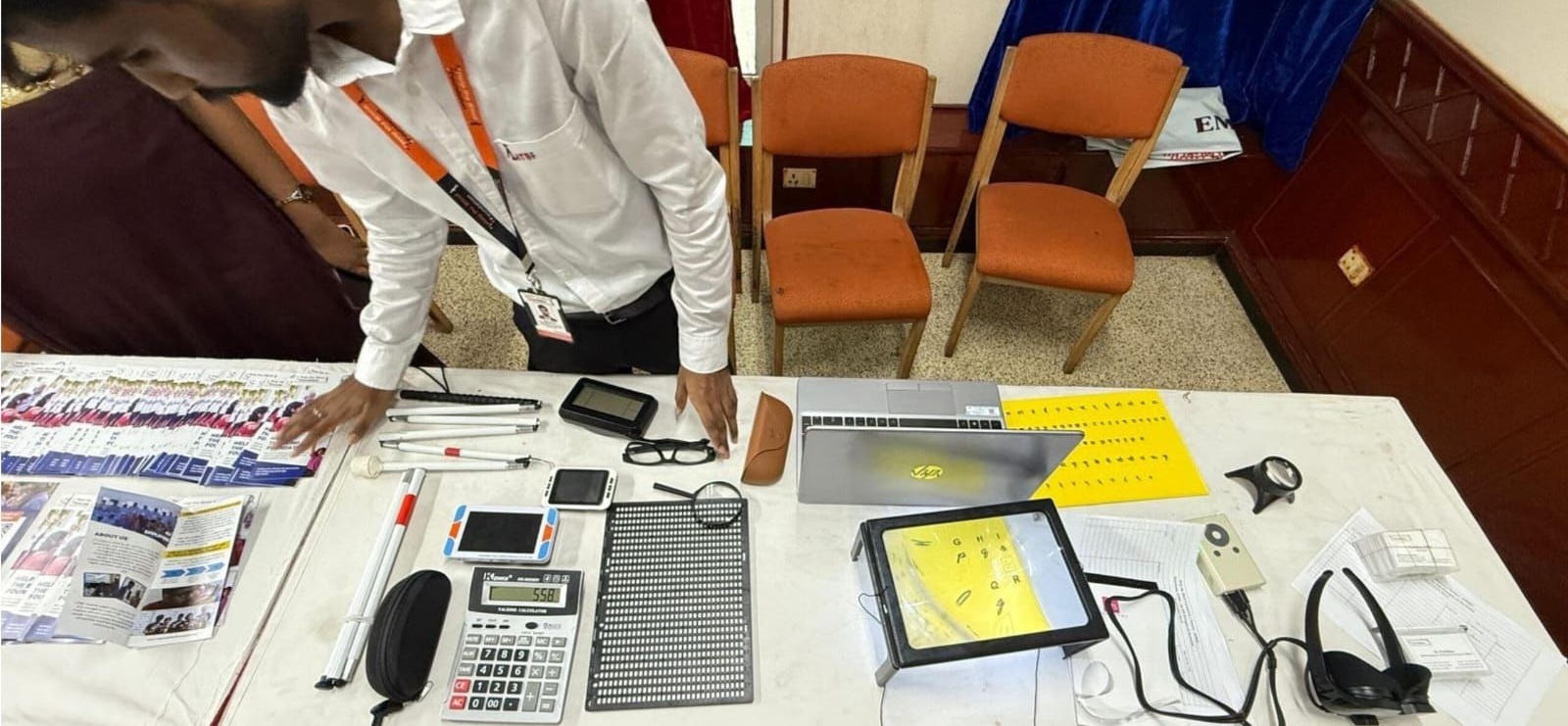 HTBF Placement and accessibility audit executive Mr.Vignesh at a stall displaying various assistive devices for visually impaired individuals. On the table there are white canes,Braille sheets, a large calculator, magnifiers, electronic devices, a laptop, brochures, and yellow tactile learning sheets.