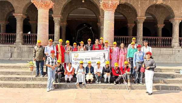 Visually impaired students from Indore pose together wearing yellow caps during a heritage walk at a historic site.