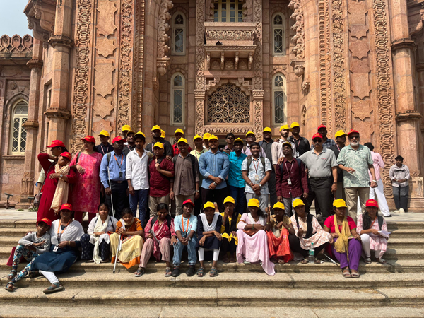 Visually impaired students and volunteers from Chennai pose together wearing yellow and red caps during a heritage walk.