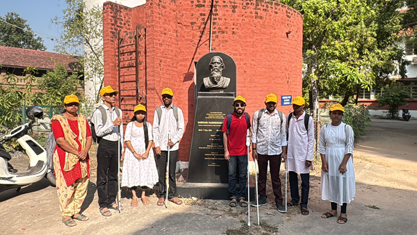 Visually impaired students wearing yellow caps stand together beside a statue of Rabindranath Tagore during a heritage walk.