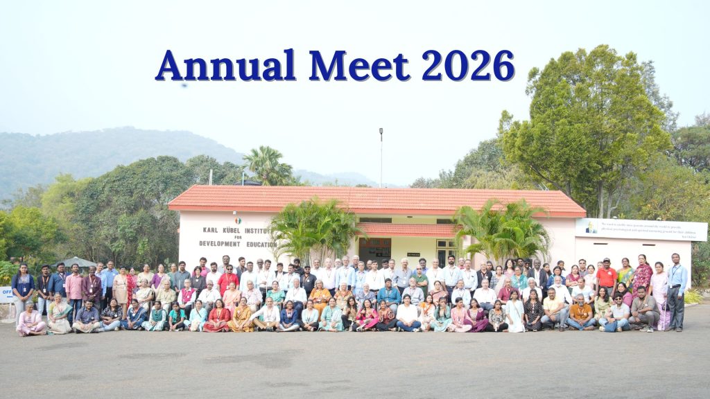 A group photo of HTBF trustees, staff, trainers, volunteers, entrepreneurs, and community members gathered outside the Karl Kübel Institute for Development Education building. The entire group is arranged in rows in front of the campus building with palm trees on either side, rolling hills in the background, and the title “Annual Meet 2026” displayed at the top of the image