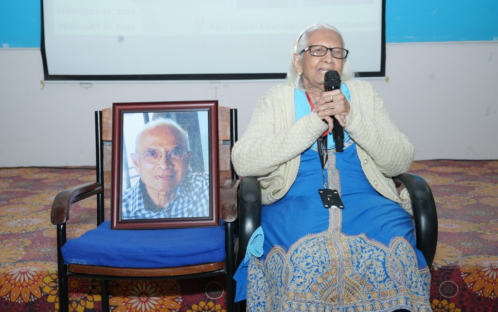 Mrs. Niti Patel sits on stage holding a microphone and addressing the audience during her inaugural speech, with a framed portrait of Mr. D.K. Patel placed beside her.