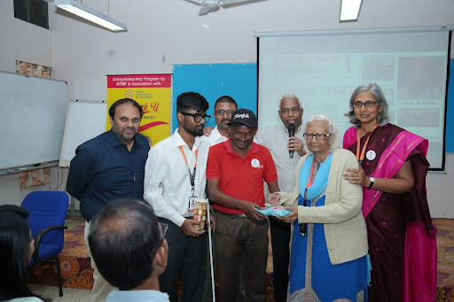 Niti Patel purchases products from a visually impaired entrepreneur at the Annual Meet 2026, as trustees and team members stand beside them.
